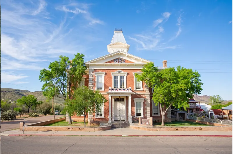 Tombstone Courthouse State Historic Park — Tombstone, Arizona The two-story red-brick 1882 Tombstone Courthouse with its white cupola, framed by green trees and desert mountains under a blue sky.